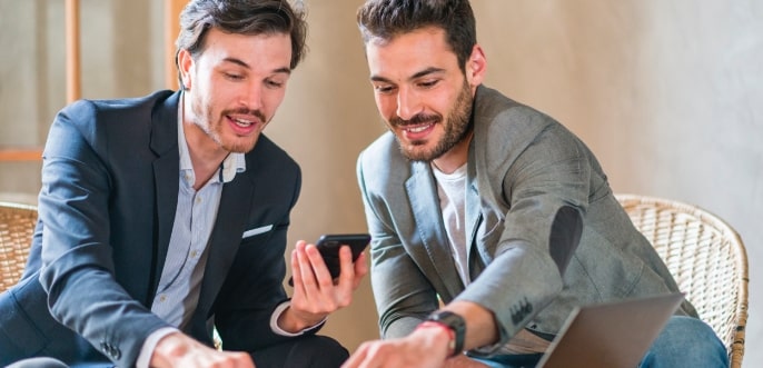 Zwei junge Männer mit Anzug unterhalten sich lebhaft und zeigen auf Ihren Laptop Bildschirm. Sie freuen sich offenkundig über Ihre Entscheidungen.