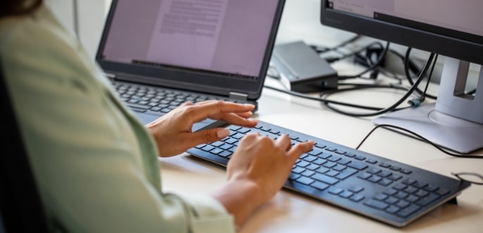 Eine Frau sitzt an Ihrem Computer und bedient die Tastatur um einen Text zu schreiben. Sie hat zur besseren Übersicht 2 Bildschirme auf ihrem Schreibtisch.